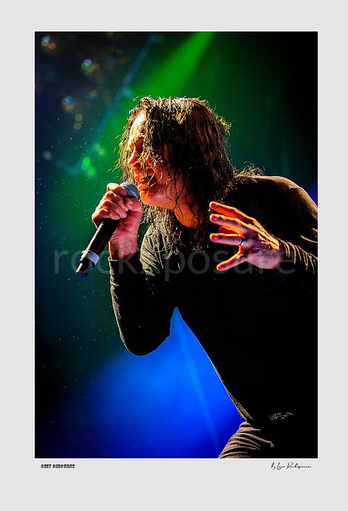 Ozzy Osbourne of Black Sabbath singing into a microphone with expressive hand gesture under dramatic green and blue stage lighting