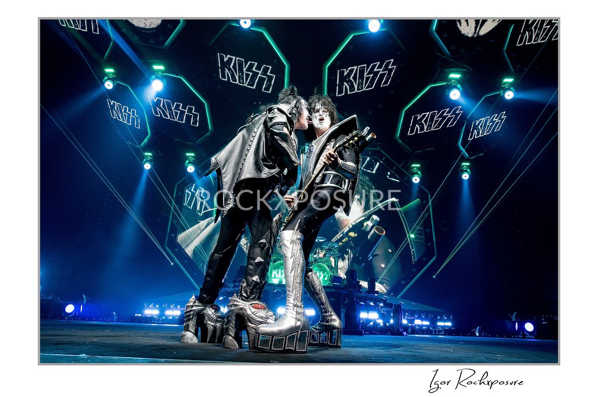 Gene Simmons of KISS leaning forward to lick Tommy Thayer’s face while both perform with bass and guitar under bright blue arena stage lighting with KISS logo stage screens behind them