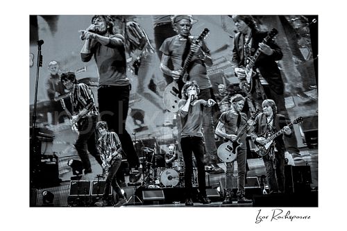 Horizontal black and white image of Ronnie Wood, Mick Jagger, Charlie Watts, Keith Richards and Mick Taylor of The Rolling Stones performing on stage with a giant backdrop screen