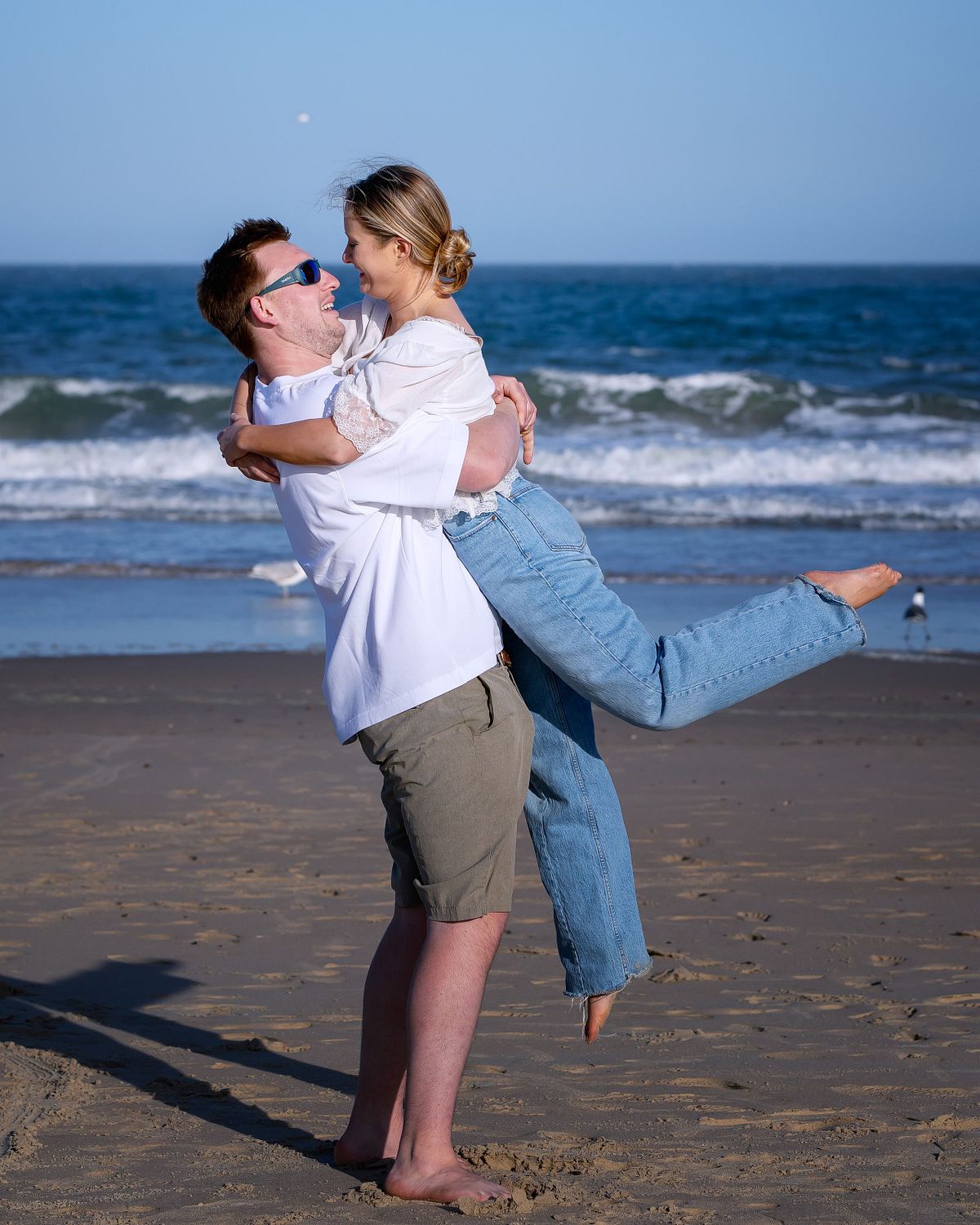 groom picking up the bride on the beach during engagement session at indian river inlet, sussex county de. she is kicking her left leg out and they are both smiling at each other