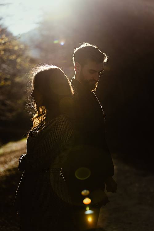 Coppia abbracciata in controluce nel bosco marchigiano, sole che filtra tra gli alberi e silhouette romantica.